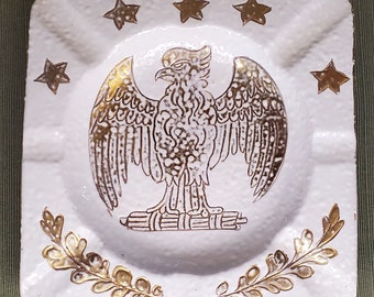 May include: White ceramic ashtray with a gold eagle design and five gold stars around the eagle. The eagle is standing on a bundle of sticks and is surrounded by two gold laurel wreaths.