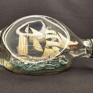 May include: A miniature sailing ship model encased in a clear glass bottle. The ship features detailed sails and is set against a blue backdrop, with green waves at the base. The bottle has a gold-coloured cap.