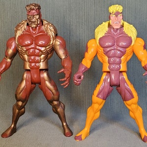 以下が含まれることがあります： 2体の筋肉質な男性キャラクターのアクションフィギュア。1体は赤と金、もう1体はオレンジと紫です。どちらのフィギュアも顔の造形が細かく、立っています。このフィギュアは、おそらく漫画やアニメシリーズからのものです。