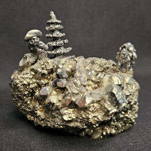 May include: A detailed pyrite sculpture featuring miniature figures and a stylized tree. The figures appear to be engaged in an outdoor scene, set atop a textured, crystalline base. The sculpture has a metallic silver and gold color.