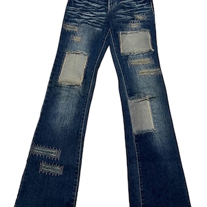 Può includere: Jeans di denim blu scuro con gamba a zampa. I jeans presentano dettagli strappati e toppe in vari punti. Le toppe sono di una tonalità di blu più chiara. Classico design a cinque tasche.