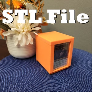 Può includere: Una scatola arancione stampata in 3D con una finestra in plastica trasparente, che mostra una carta di Magic: The Gathering. La carta è intitolata "Solemn Simulacrum" e fa parte del set "Modern Horizons 2". La scatola è appoggiata su un sottopentola circolare blu.