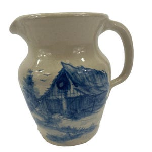 May include: A beige ceramic pitcher with a blue barn scene painted on the front. The pitcher has a curved handle and a spout for pouring. The design is reminiscent of vintage farmhouse decor.