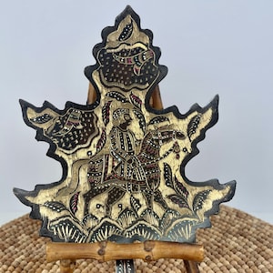 May include: A black metal leaf-shaped wall hanging with gold and colored accents depicting a person riding a horse. The wall hanging is displayed on a small bamboo easel.