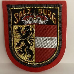 May include: Embroidered patch featuring the Salzburg coat of arms. The patch has a red border and a shield design with the word "SALZBURG" arched across the top. The shield is divided into sections with a gold lion, red and white stripes.