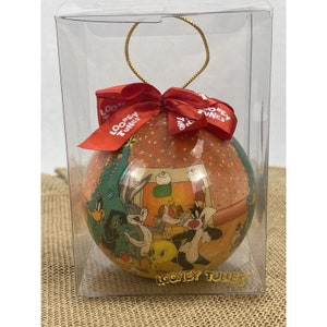 May include: A red and gold Christmas ornament featuring a scene of Bugs Bunny, Tweety Bird, Sylvester the Cat, and Daffy Duck celebrating Christmas. The ornament is in a clear plastic box with a red bow.