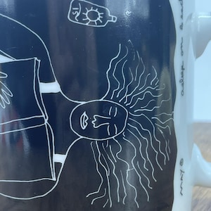 May include: A black ceramic mug with white line art. The design features a stylized face with sun-like rays and the text "asleep on beach". The mug has a white handle and a glossy finish.