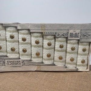 May include: A set of eight white and gold striped coasters with gold buttons. The coasters are packaged in a white box with the text "COASTER CLASSICS" and "BY HI-JAC" printed on it.