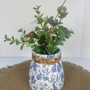 May include: A white ceramic vase with a blue and white rabbit pattern and gold trim. The vase is filled with a green leafy plant.