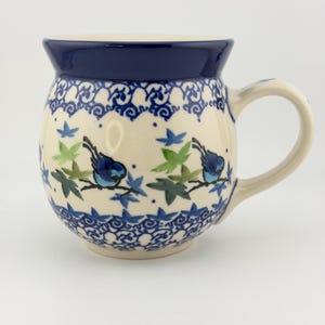 May include: A round ceramic mug with a cream-colored base and a dark blue rim. The body features a repeating pattern of small blue birds perched on branches, interspersed with green and blue leaves, framed by decorative blue scrollwork borders.