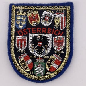 May include: A blue embroidered patch with a gold border and the word "ÖSTERREICH" in red. The patch features multiple crests and emblems, including a central double-headed eagle and various regional coats of arms.