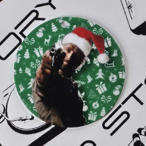 May include: A round Christmas ornament featuring a man wearing a Santa hat and pointing a gun. The ornament has a green background with white Christmas-themed patterns. The ornament is hanging from a white string.