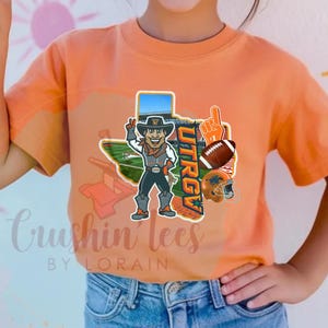 May include: Orange t-shirt featuring a cartoon cowboy with a Texas outline, football, and helmet graphics. The shirt includes the text "UTRGV" and "Crushin' Tees by Lorain".
