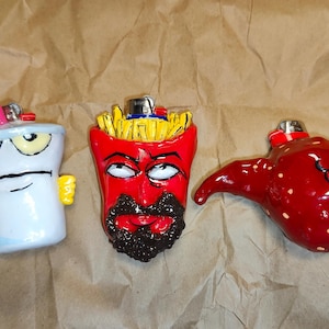 May include: Three custom lighter cases featuring sculpted characters. The left is a light blue cup with a pink straw, the center is a red container of yellow french fries with a black beard, and the right is a red, textured, blob-like character.