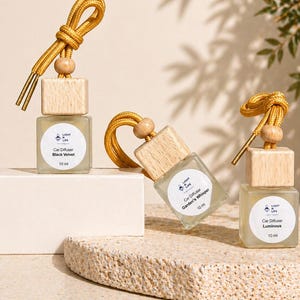 May include: Three car air fresheners with wooden tops and gold cords. Each glass bottle has a label with the text "Car Diffuser" and a scent name: "Black Velvet", "Garden's Whisper", and "Luminous". Each bottle contains 10 ml.