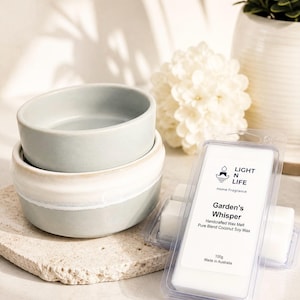 May include: Two small, pale blue and white ceramic bowls stacked on a stone slab. Beside them are two packages of wax melts, labelled "Garden's Whisper" with "LIGHT N LIFE" branding. A white flower and a potted plant are in the background.