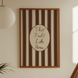 May include: A framed artwork with a brown and white striped design. The artwork has an oval shape in the centre with the handwritten text "This Feels Like Home". The frame is brown, and the piece is hanging on a white wall, near a staircase.