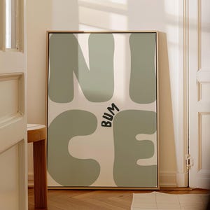 Könnte beinhalten: Ein gerahmter Kunstdruck mit dem Wort "NICE" in großen, abgerundeten, salbeigrünen Buchstaben und dem Wort "BUM" in schwarzer, stilisierter Schrift. Der Hintergrund ist cremefarben. Der Rahmen ist goldfarben.