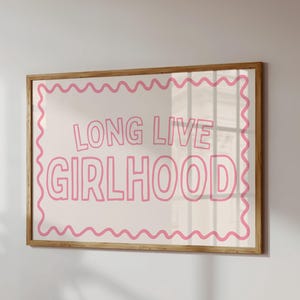 Pode incluir: Uma impressão emoldurada com as palavras "LONG LIVE GIRLHOOD" em rosa, texto delineado sobre um fundo creme. A impressão tem uma borda ondulada rosa e uma moldura de madeira natural. A obra de arte está pendurada em uma parede branca.