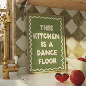 Puede incluir: Una escena de cocina con un letrero que dice "THIS KITCHEN IS A DANCE FLOOR". La encimera muestra un fregadero, un grifo dorado y granadas. Una estantería de madera encima contiene tazas y vasos. El fondo incluye una ventana y azulejos estampados.