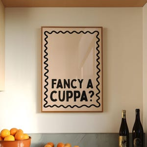 May include: A kitchen scene with a framed print that reads "FANCY A CUPPA?" in black text. A terracotta bowl filled with oranges sits on a gray countertop, along with several loose oranges. Two wine bottles are also on the counter.
