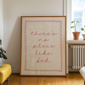 以下が含まれることがあります： ピンク色の筆記体で「there's no place like bed」と書かれた額入りのアートプリント。プリントはクリーム色の背景とピンク色のスカラップボーダーです。フレームは明るい茶色です。プリントは白い壁に立てかけられています。