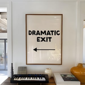 May include: A framed wall art print featuring the black text "DRAMATIC EXIT" above a black left-pointing arrow on a cream background. The print is housed in a simple brown wooden frame and is displayed on a rustic wooden console table.