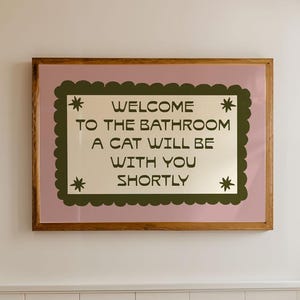 Puede incluir: Un interior de baño con una bañera blanca, un taburete de madera con artículos de tocador y un letrero enmarcado. El letrero dice "WELCOME TO THE BATHROOM A CAT WILL BE WITH YOU SHORTLY" en texto verde sobre un fondo crema, enmarcado en rosa y madera.