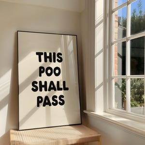 May include: A rectangular wall art print in a thin black frame featuring the bold black text THIS POO SHALL PASS centered on a cream-colored background. The print is displayed in a vertical orientation with clean, rounded sans-serif typography.