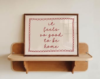 Könnte beinhalten: Ein gerahmter Druck mit dem roten Text "it feels so good to be home" in Schreibschrift. Der Text ist von einem roten, wellenförmigen rechteckigen Rand auf cremefarbenem Hintergrund umgeben und in einem dunkelbraunen Holzrahmen.