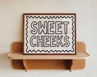 May include: A rectangular print in a dark wood frame featuring the text SWEET CHEEKS in bold black capital letters. The text is enclosed within a black wavy border on a white background, displayed on a light wood wall-mounted shelf.