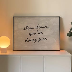 May include: A framed print with the words "slow down, you're doing fine" is displayed above a white cabinet. A spherical lamp emits a warm glow. A large green plant is visible on the right.