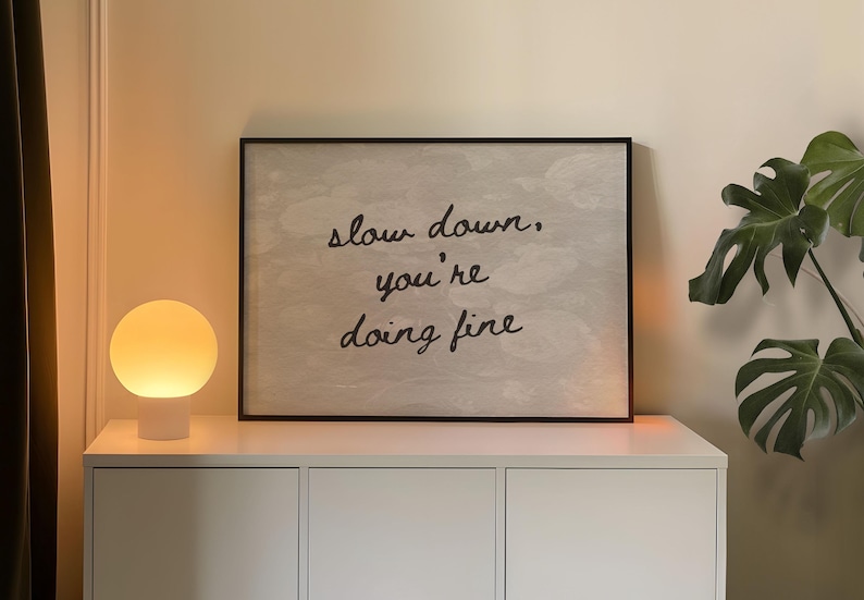 May include: A framed print with the words "slow down, you're doing fine" is displayed above a white cabinet. A spherical lamp emits a warm glow. A large green plant is visible on the right.