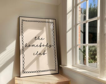 May include: A rectangular wall art print in a thin black frame featuring a cream-colored background with a hand-drawn scalloped border in black ink. The center of the print displays the black cursive text that reads the homebody club.