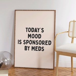 May include: A framed print with a light pink background and black text that reads "TODAY'S MOOD IS SPONSORED BY MEDS". The print is hanging on a white wall next to a wooden chair with a white cushion.