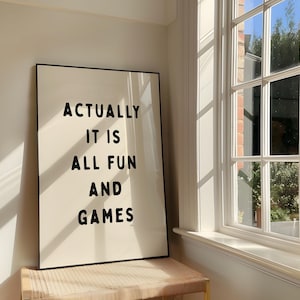 May include: A framed art print with the text "ACTUALLY IT IS ALL FUN AND GAMES" in black block letters. The print is leaning against a wall next to a window with sunlight streaming through.