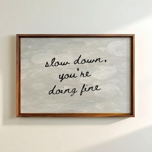 May include: A framed print with the words "slow down, you're doing fine" in black script. The background is a light grey with a subtle pattern. The frame is a dark brown wood.