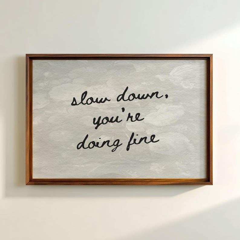 May include: A framed print with the words "slow down, you're doing fine" in black script. The background is a light grey with a subtle pattern. The frame is a dark brown wood.
