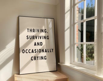 May include: A framed print with the text "Thriving, Surviving and Occasionally Crying" in black letters on a cream background. The print is displayed near a window with sunlight streaming through.