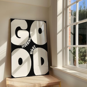 May include: A rectangular black and white art print featuring the words "GOOD MORNING NIGHT". The artwork is displayed indoors, near a window. The print has large, bold letters. The artwork is mounted on a wall, with sunlight streaming through the window.