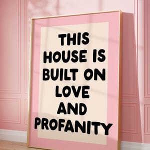 May include: A framed print with a pink background and a white rectangle in the center. The text "THIS HOUSE IS BUILT ON LOVE AND PROFANITY" is printed in black on the white rectangle.