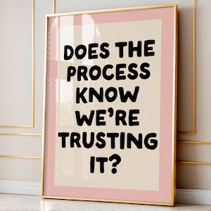 Op de afbeelding: Ingelijste kunstdruk met de tekst "DOES THE PROCESS KNOW WE'RE TRUSTING IT?" in vette zwarte letters. De print heeft een lichtbeige achtergrond met een roze rand en een gouden lijst. Het kunstwerk wordt aan een muur getoond.