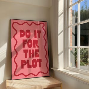 May include: A rectangular wall art print in a black frame featuring a pink background with a wavy red border. The text DO IT FOR THE PLOT is printed in bold, red, sans-serif capital letters centered on the pink field within the wavy border.