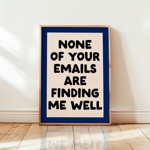 May include: A framed art print with a navy blue border and a beige background. Bold black text reads "NONE OF YOUR EMAILS ARE FINDING ME WELL". The artwork is displayed on a wooden floor against a white wall.