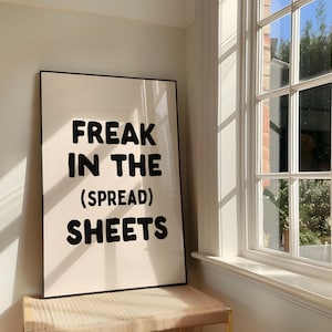 Può includere: Un'opera d'arte rettangolare incorniciata con uno sfondo color crema e un testo nero in grassetto, arrotondato e senza grazie che recita: FREAK IN THE (SPREAD) SHEETS. La stampa è racchiusa in una sottile cornice nera ed esposta alla luce.