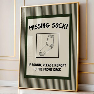 Op de afbeelding: Een ingelijste verticale poster met een beige achtergrond en een donkergroene gestreepte rand. De tekst luidt MISSING SOCK! en IF FOUND, PLEASE REPORT TO THE FRONT DESK. Een zwart-witte lijntekening van een enkele sok staat centraal in een vierkant.