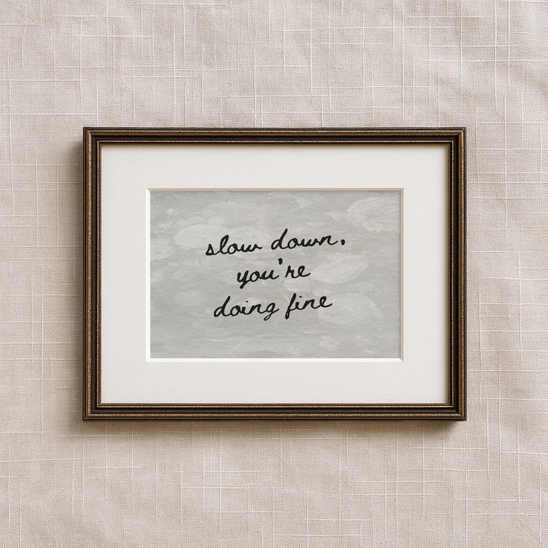May include: A framed print featuring the text "slow down, you're doing fine" in black script. The print has a white mount and a dark brown frame. The background is a light grey, textured design.