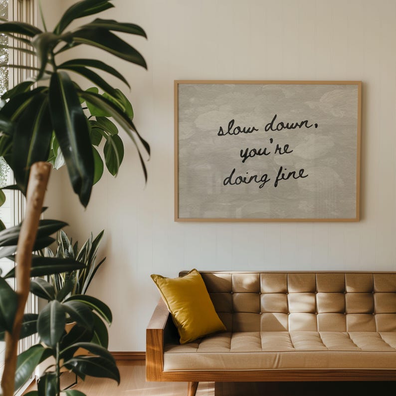 May include: A wooden framed print with the words "slow down, you're doing fine" is displayed above a tan leather sofa with a yellow cushion. A large potted plant is in the foreground. The room has a warm and inviting atmosphere.