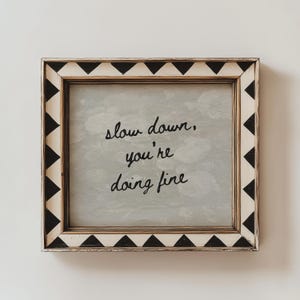 May include: Square framed art with the message "slow down, you're doing fine" in black script. The frame is white with black triangle accents. The background has a soft, cloudy appearance.