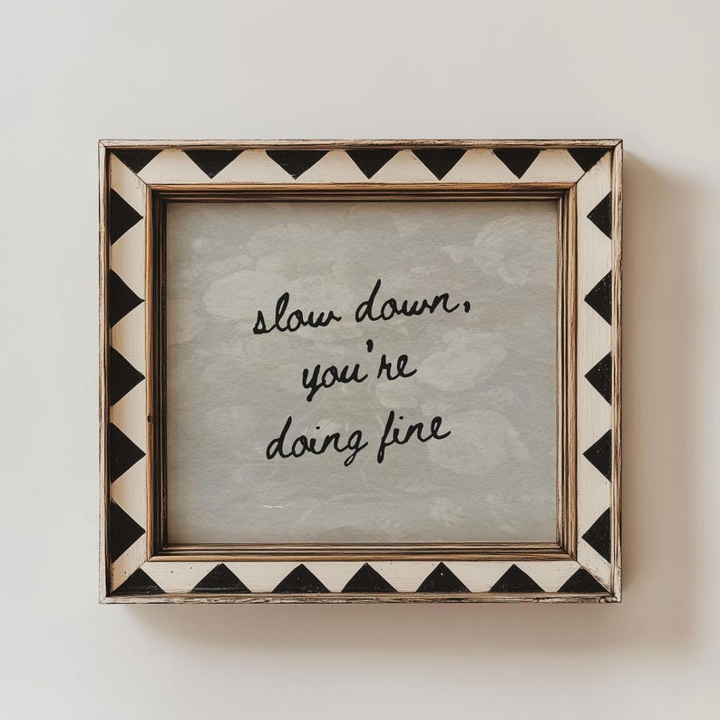 May include: Square framed art with the message "slow down, you're doing fine" in black script. The frame is white with black triangle accents. The background has a soft, cloudy appearance.